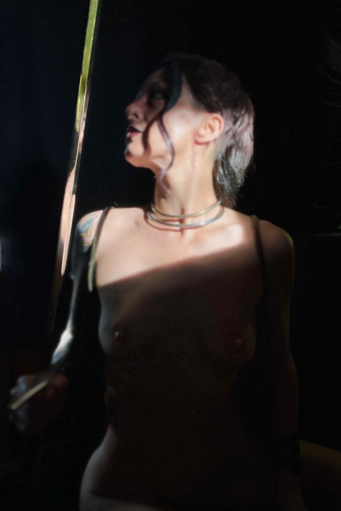 Nat is seen from her hips to her face-

she is heavily shadowed- illuminated only by slivers of light from behind her and a large diagonal slash of light across her lower face and décolletage. Her face tilted up and turned to frame left looks at the sword she holds in her hand at waist height. The sharp blade catches all available light making the steel glow. Her green velvet dress is split down the front and is currently laid open to reveal the entirety of her torso- the limited light creating a defined halo around her body frame left. Her only adornments her double stack of locked collars. 
