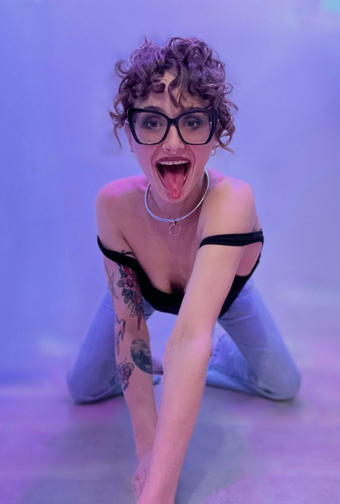 we see that against a pastel blue and pink and white background-softly washed mixing the colors-that is on all fours with her shoulders on face center frame. She wears pale wash blue jeans and a black ribbed sleeveless bodysuit, the straps pulled off her shoulders.
Her natural curly brown hair is pinned high on her head and falls in Ringletts around her face-her eyes are mischievous from behind thick black plastic butterfly frame glasses. She wears a steel locked collar with a gold O-ring. 
she is grinning at the camera and sticks at her tongue as she appears to prowl towards the viewer. 