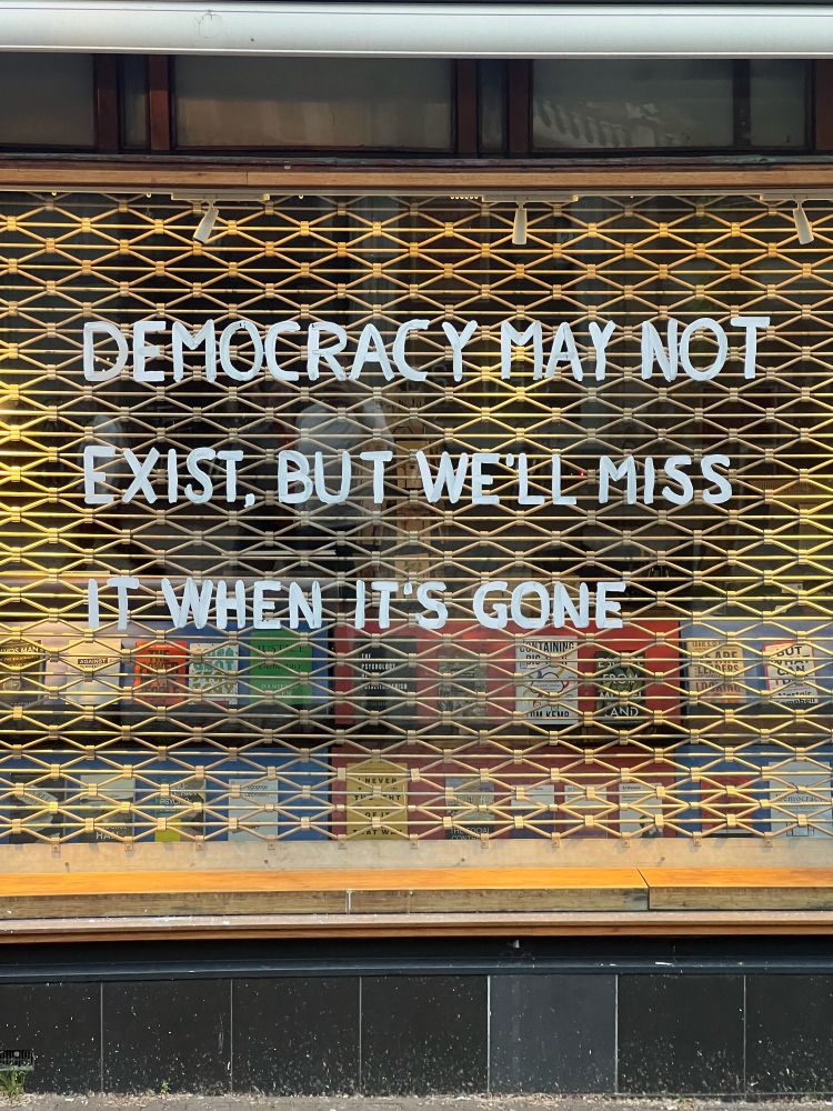 "DEMOCRACY MAY NOT EXIST, BUT WE'LL MISS IT WHEN IT'S GONE" painted in block letters on the window of a closed bookstore in Amsterdam, NL