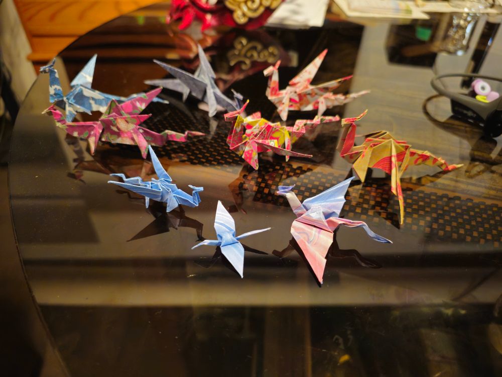 An array of 8 origami dragons of varying designs, and one origami crane. They are all on a dark reflective surface. I failed to hide all of the mess on other parts of the table too.