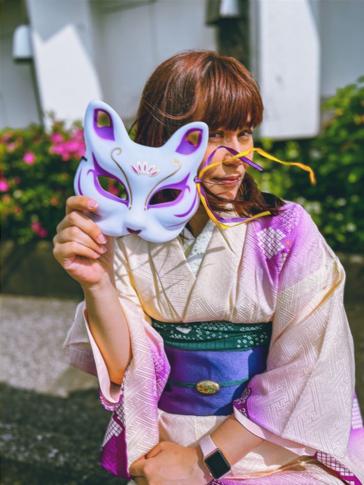 The gorgeous Ishikawa Sachiko wearing a beautifully patterned kimono with shades of purple, white, and green poses while holding a purple and white fox mask with golden details to the side of their face. Their hair frames their face as they glance toward the camera, and the background features a mix of greenery and an urban structure.