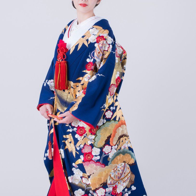 The image shows a bride wearing an iro-uchikake, a colorful Japanese bridal kimono. The kimono is a rich blue color with elaborate embroidery featuring floral motifs, including plum blossoms and bamboo leaves, along with gold accents. The bride also wears a red tassel accessory on her chest, adding a bright contrast to the blue fabric. The design and vibrant colors of the kimono symbolize happiness, prosperity, and good fortune. The bride holds a small red fan, enhancing the traditional bridal ensemble.