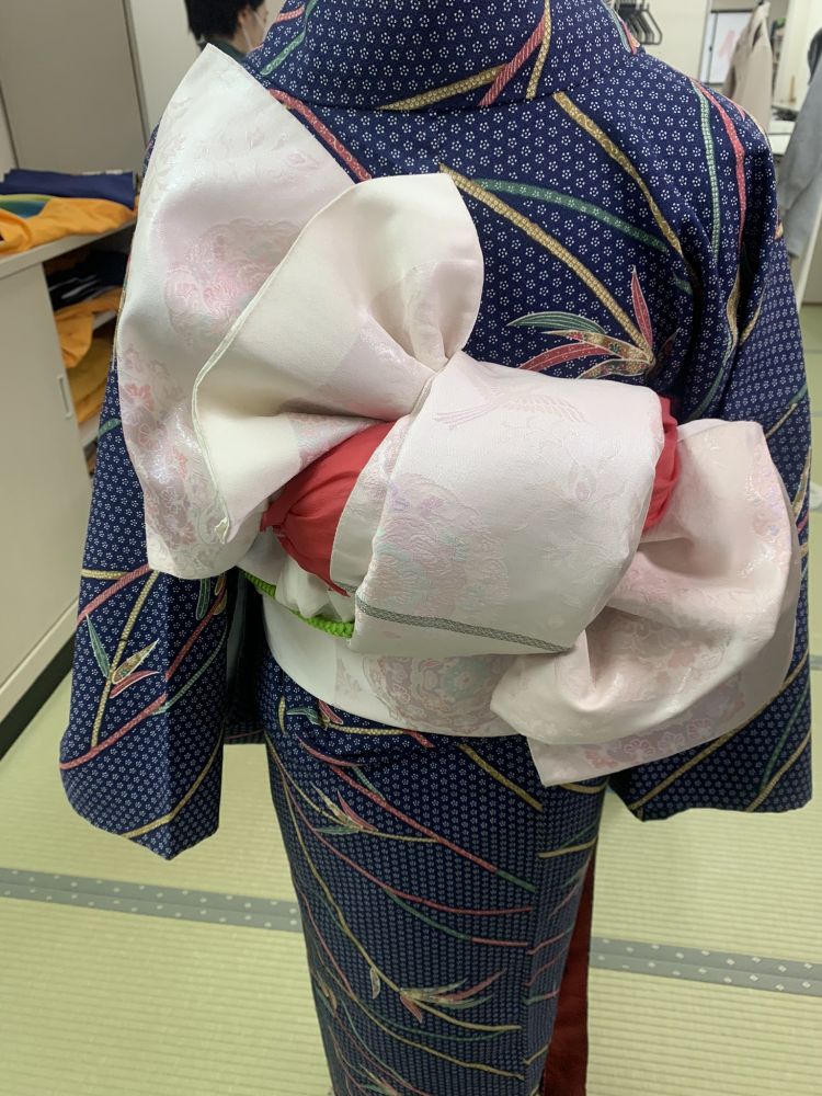 Close-up view of a traditional kimono with a pink and white obi tied in an elaborate style. The kimono features a blue fabric with intricate patterns. The background shows a tatami mat. It's from kimono school practice.