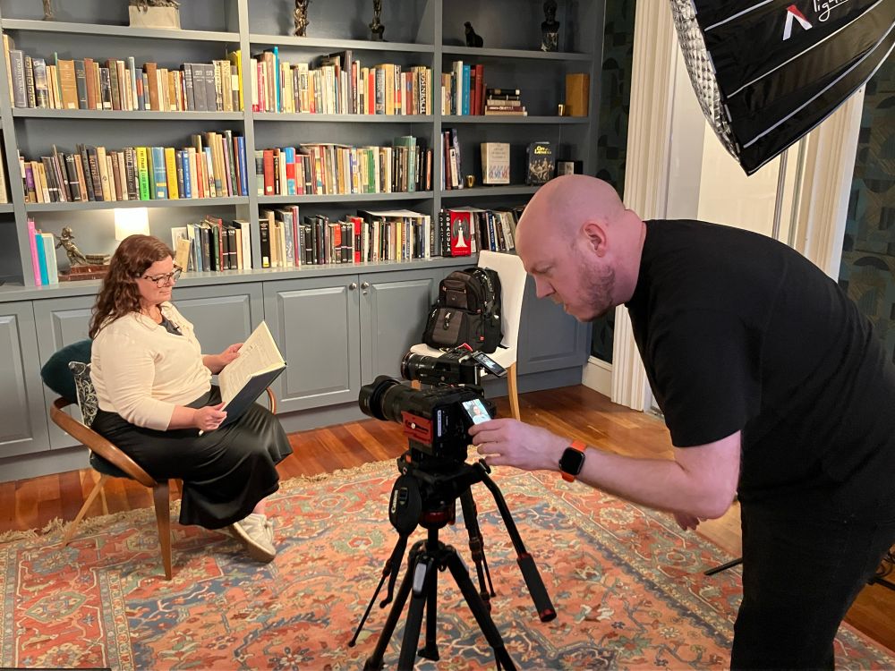 Emilie Parker sits reading from a large book before two cameras on tripods and lighting equipment 