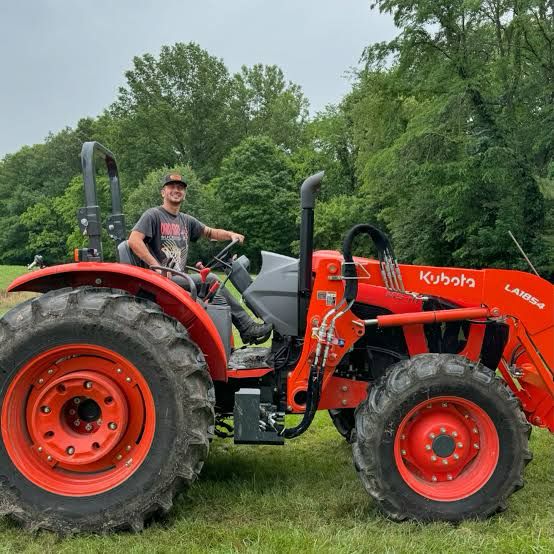 a man is sitting in the driver's seat of a red Kubota tractor with a front-end loader attachment. The tractor is parked in a grassy field with a line of trees in the background. The man is wearing a t-shirt, shorts, and a baseball cap.