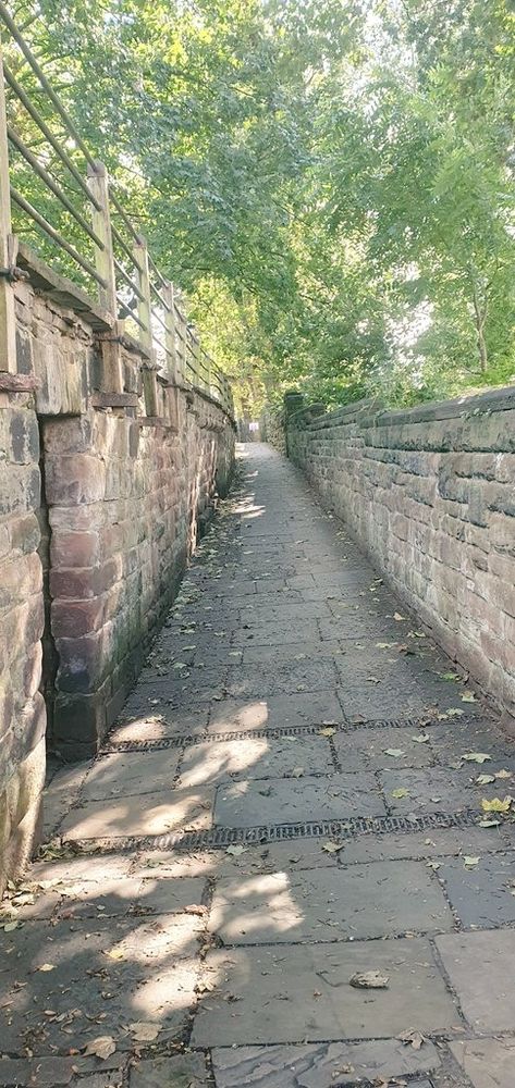 Sandstone former defensive town wall with small gate on the left. Railings on left hand side. Sunny day with sunlight coming through and leafy green trees ahead