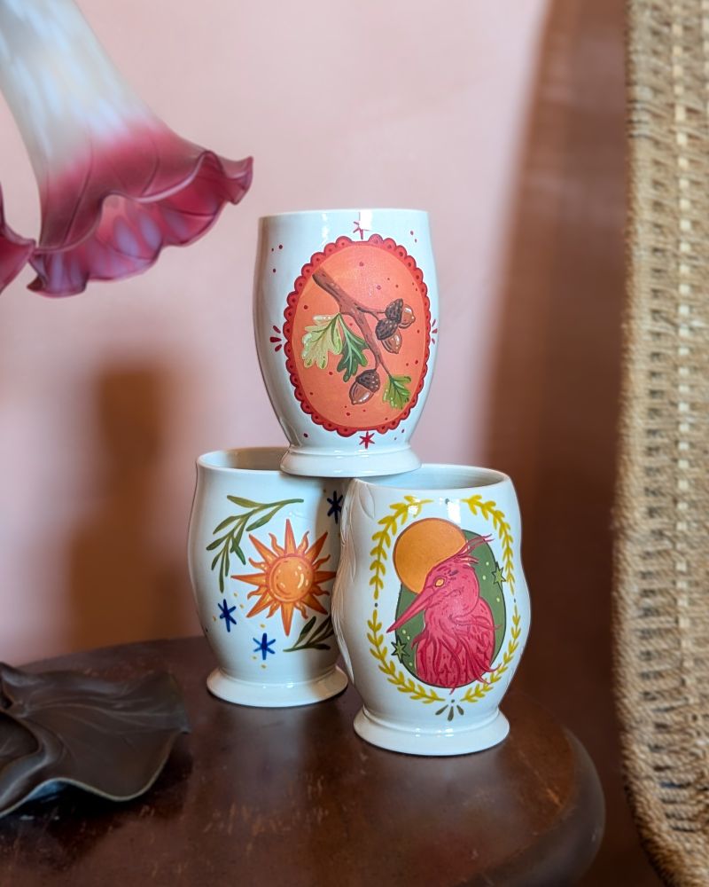 Three colorfully painted porcelain cups stacked together, with a pink heron, a sun, and an acorn design. 