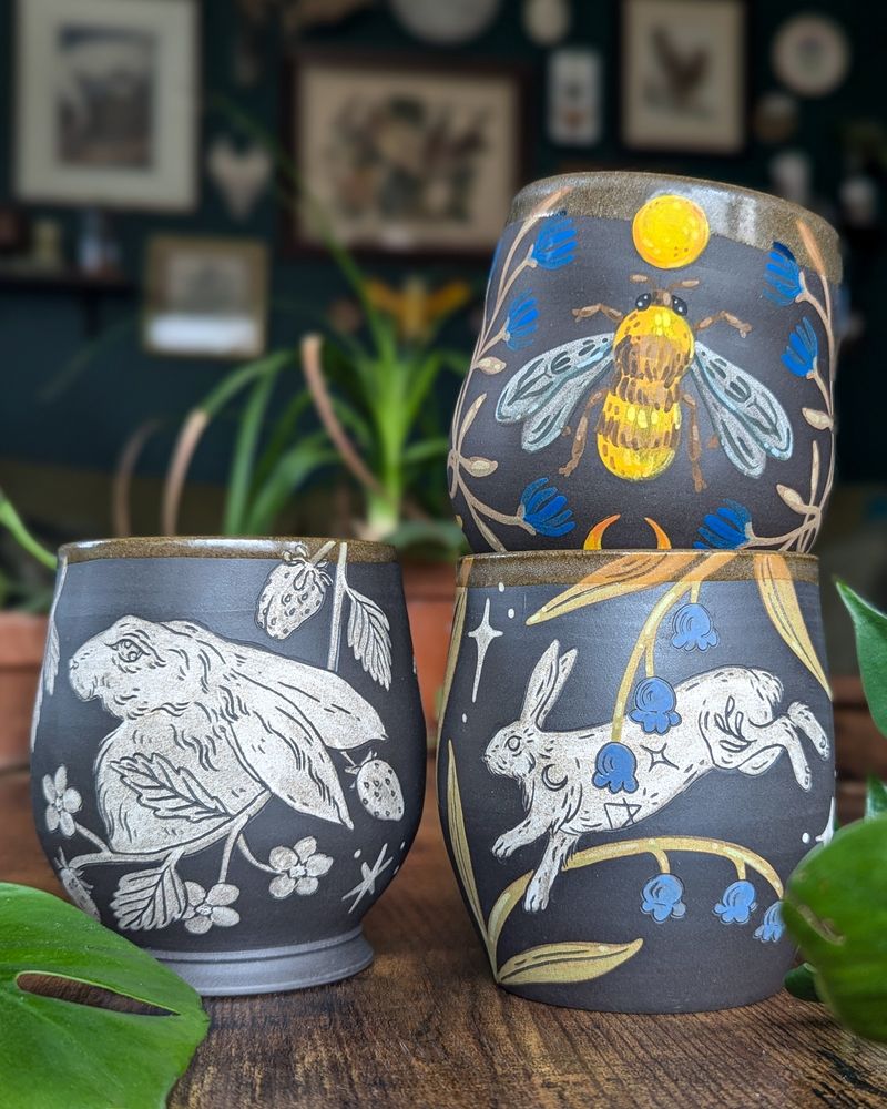 Three dark brown ceramic cups stacked together, two with rabbits, flowers and strawberries, and one with a bright yellow bee framed by blue flowers. 