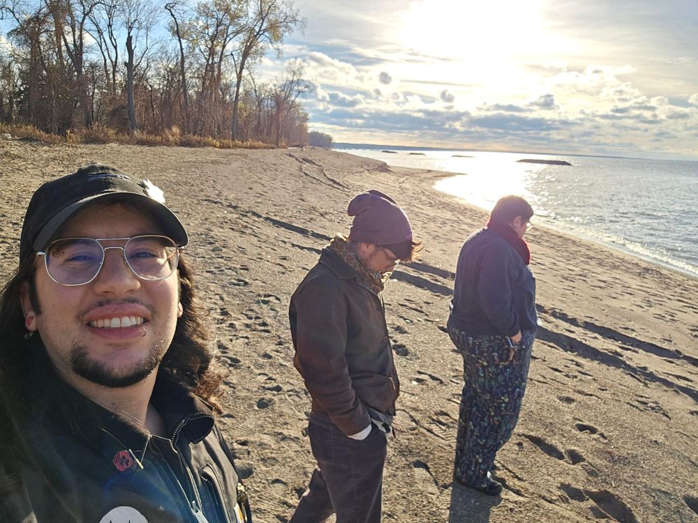 Hana taking an ussie at the beach. Hana has long dark hair, a goatee, and glasses. in the middle is Merle with his brown leather llbean coat, brown pants, and a purple beanie hat. after Merle is serendipity looking out at the sometime reflecting off the lake. there aren't a lot of leaves left on the trees and it's a little windy.