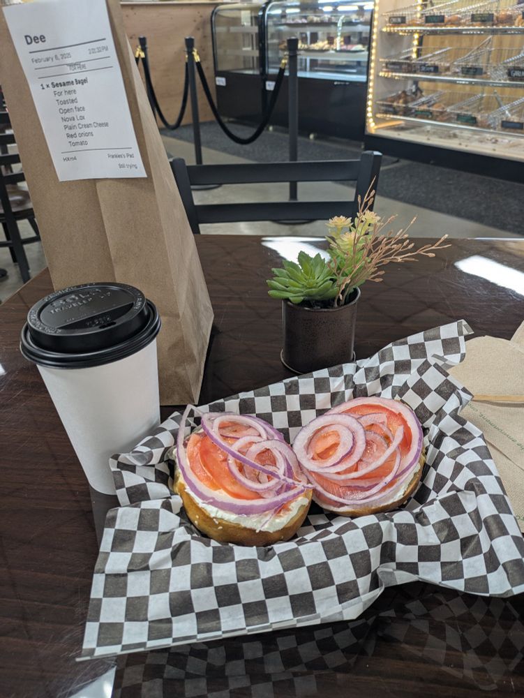 Tomato red onion lox and a schmeer at Frankie's New York Bagels of Humboldt. The bagels are nested in a black and white checkerboard paper next to a coffee and a little pot of succulents. 