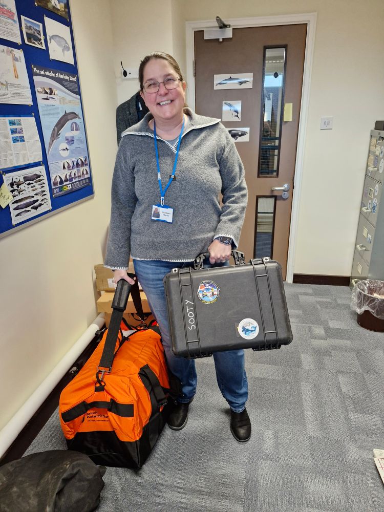 Hungry Humpbacks project manager Steph Martin, packed and ready to go. She is standing in an office holding a black peli-case and an orange kit bag. 