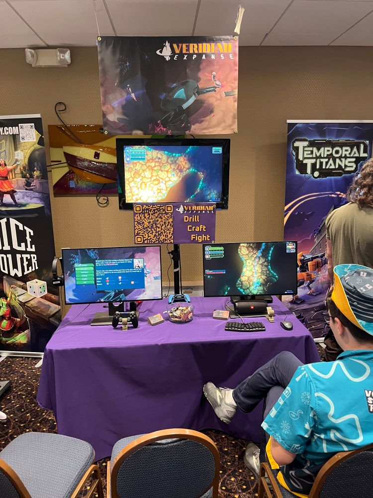 My Veridian Expanse demo booth with a banner hanging from the ceiling, a TV on a stand, and a pair of computer monitors on a purple table cloth.