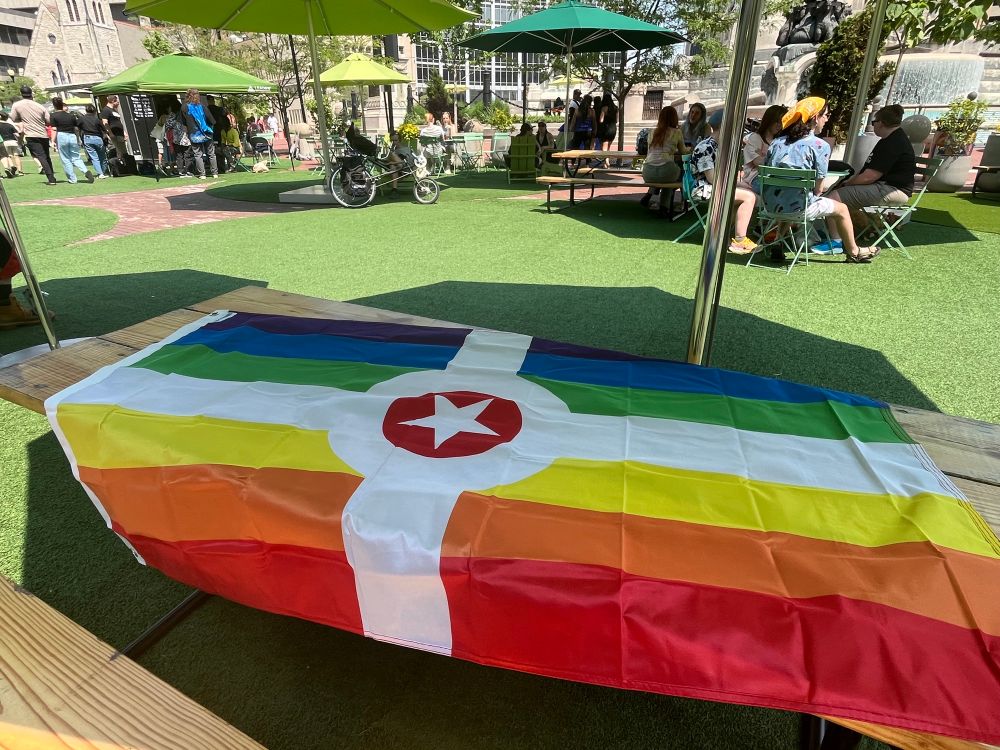 An Indianapolis city flag, with the blue replaced by a rainbow, sitting on a picnic table under an umbrella on a fake green lawn in the downtown circle.
