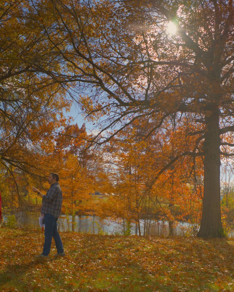 Ed speaks in front of the re-dug lake at Washington Park, a small pond now, fringed by yellow and orange foliage, as low sun shines through the trees.