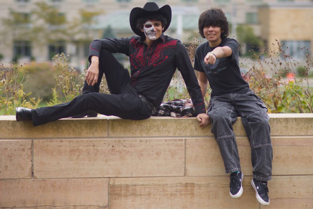 Two youth sit on a wall, one dressed as a vaquero in black, his face half made up as an elaborate skull.