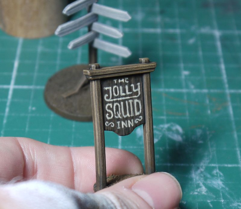wargame model. a small wooden sign. the name on it reads: 'The Jolly Squid Inn'. Maybe a wretched hive of scum and villainy, or a surprisingly class winebar.