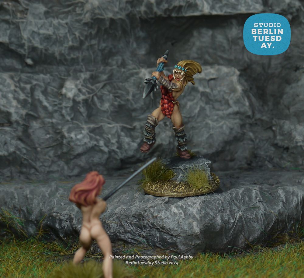 wargame models. two warrior women fight to the death. one wears dragon scale armor and holds a spear. she is leaping toward another warrior woman wearing nothing. she holds a broadsword. They fight in grassy rock surroundings.