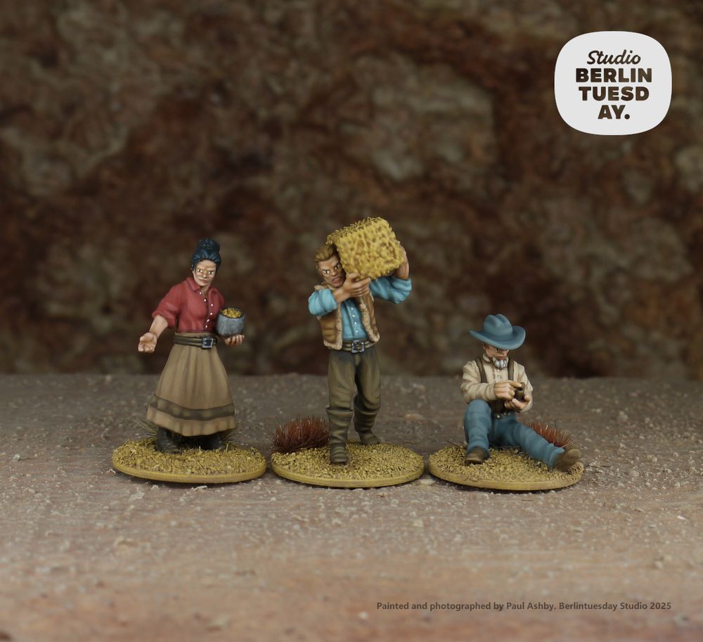 wargame models. three old west civilians and/or farmers. all wear muted colours of red blue and brown. one woman is holding a grain pan and scattering feed. one of the men has a bail of hay on one shoulder and the other man in sitting on the ground packing his pipe to smoke.