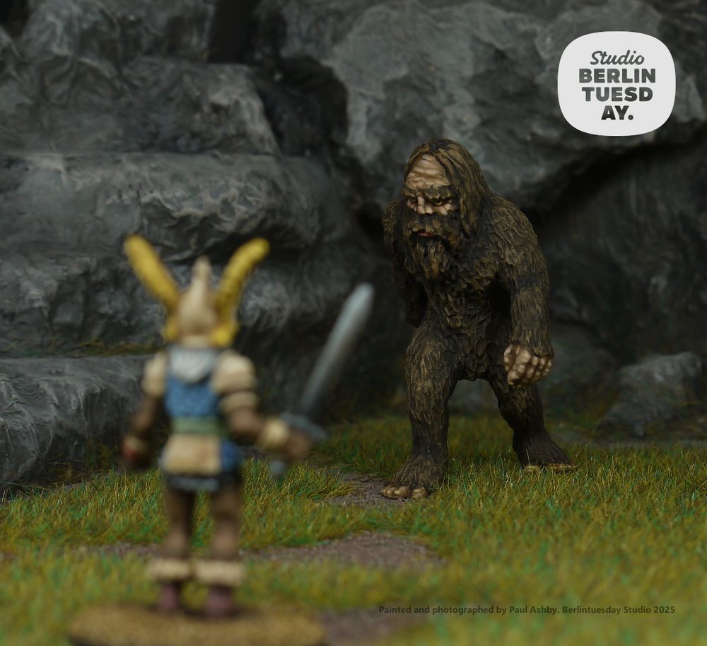 wargame model scene. in the foreground a fantasy warrior holds a sword in reddiness to take on a brown, hairy sasquach. rocky background and grass.