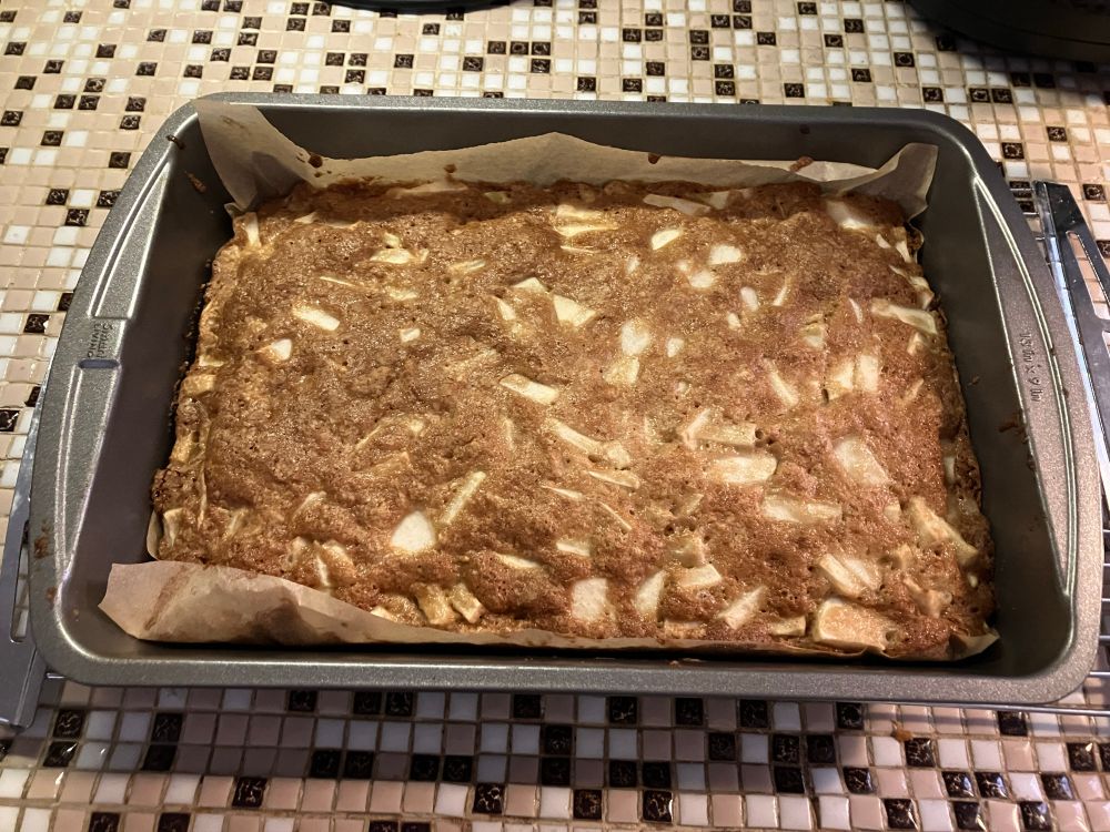 A fresh apple cake out of the oven and on a cooling rack. There are apple pieces nicely visible from the top of the cake. Parchment paper is seen from the two long sides of the rectangular pan, preventing the cake from sticking to the bottom.