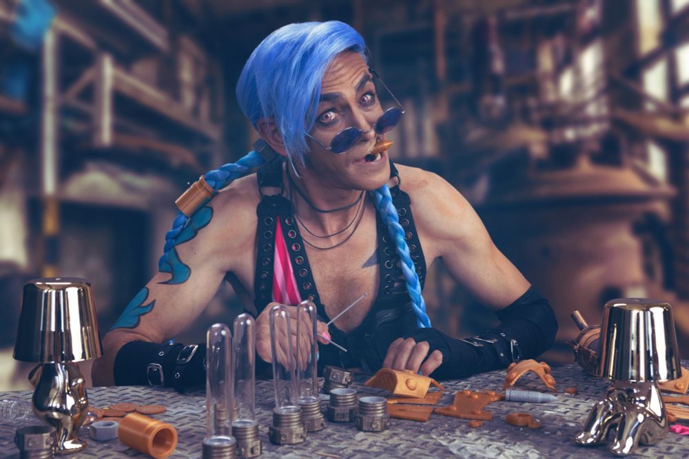 A cosplayer portraying Jinx from Arcane sits at a workbench covered in tools and spare parts