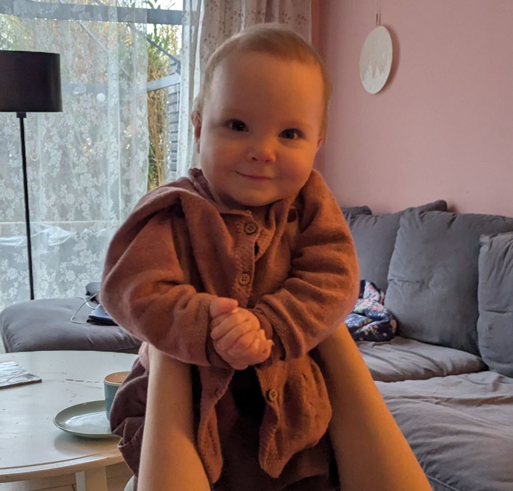 Lächelndes Kleinkind wird in Kamera gehalten und hält dabei die Hände verschränkt 