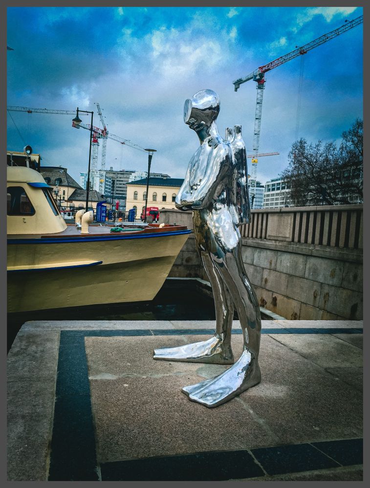 The picture shows a harbour scene in Norway in the capital city of Oslo. Prominent in the picture is a life-size statue of a frogman/diver which is metallic from head to toe and highly reflective. The frog man is on a concrete plinth in the harbour with a boat which is cream in colour behind him. In the far background there are various buildings and also tall cranes dotted around the landscape. Above there is a mottled blue cloudy sky the whole picture is in colour with a grey border surrounding it.