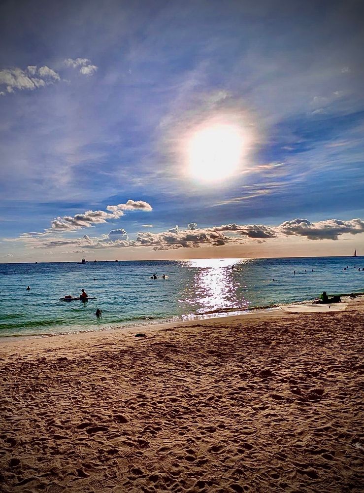 The picture shows a beat scene from a vacation in the Philippines a few years ago. There is golden sands in the foreground with the beautiful blue aqua colour of the ocean piercing the horizon. 

Above there are light clouds, a beautiful blue sky and a muted orange sun. In the water you have a few bathers, some people that are on the beach itself and in the far distance to the right there is a sailing boat. 