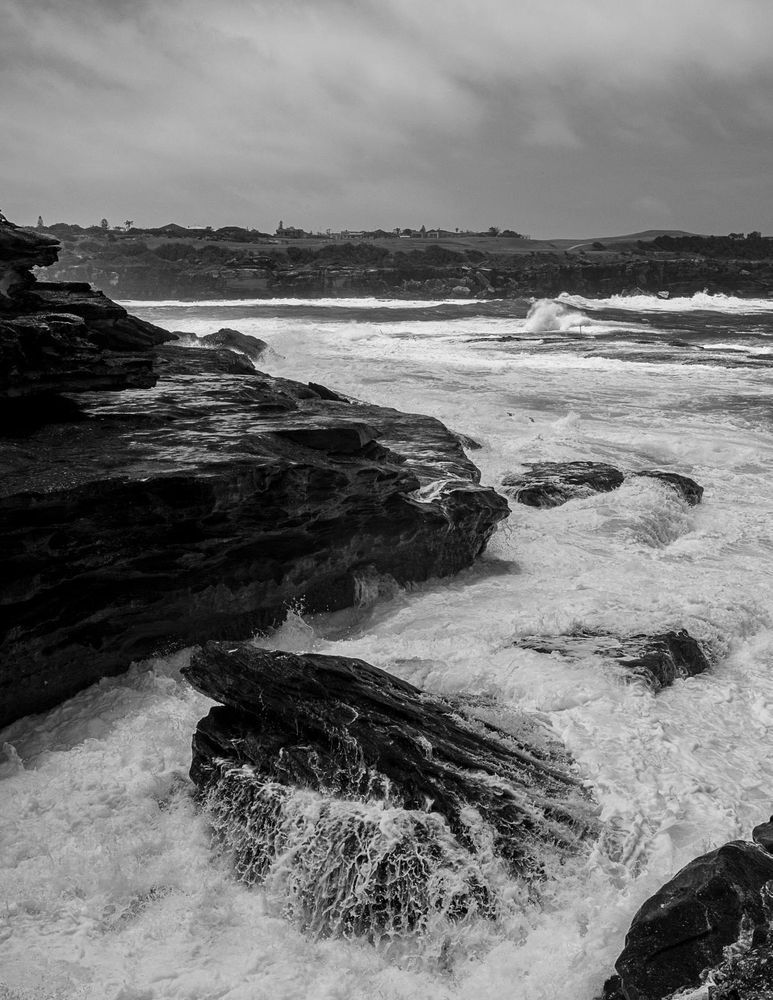 A moody black and white photo of waves breaking over coastal rocks. Lots of white foam from the swell is visible.
