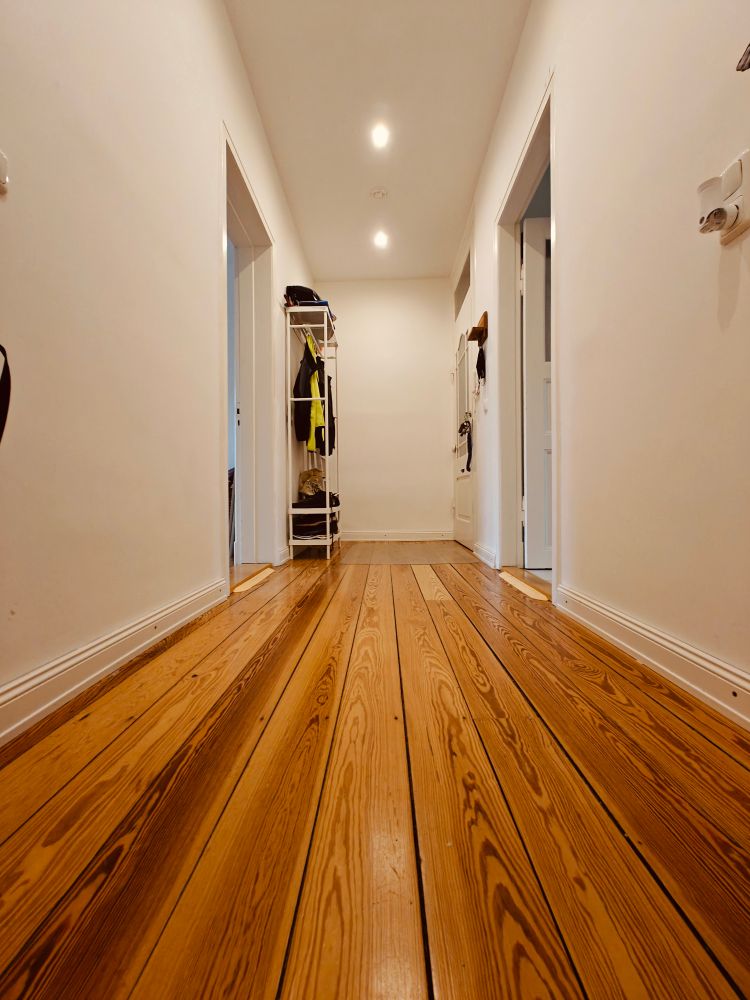 Blick auf einen nahezu leeren Flur. Ein markanter Holzfußboden, hinten links ist gegenüber der Eingangstür eine Garderobe zu sehen.