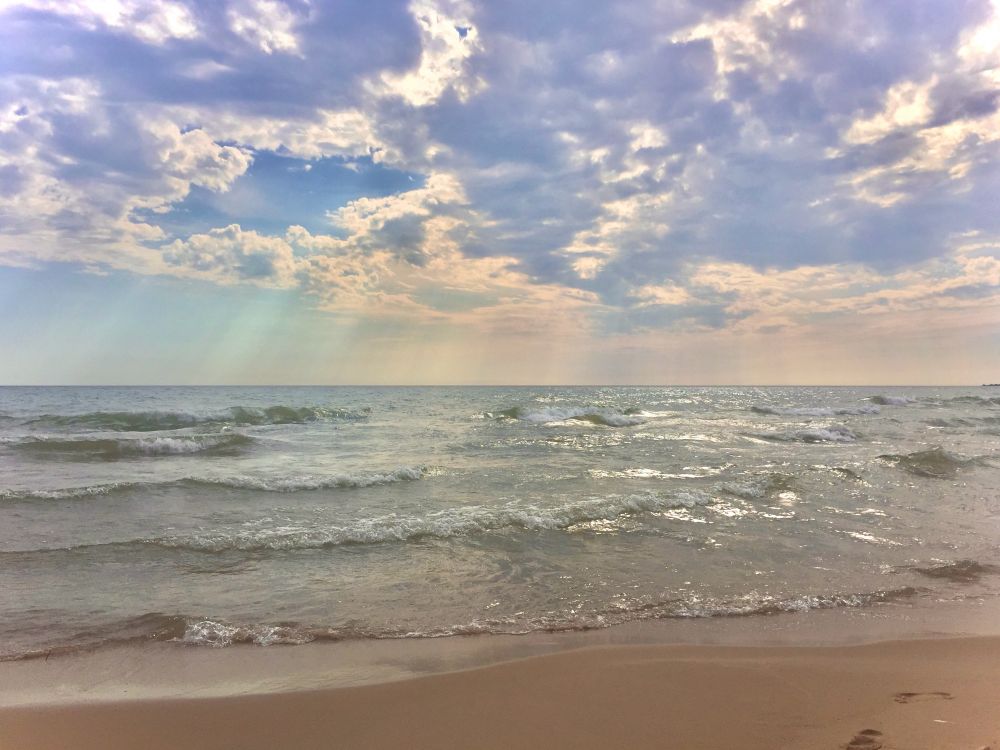 Photo of a Lake Michigan shoreline. Sunbeams filter through scattered clouds onto lively waves rolling into a beach of clean brown sand. A pair of footprints is visible in the wet sand. The sunlight shimmers from blue to green to gold across the water, which fills the horizon, no other land in sight, as far as the eye can see.