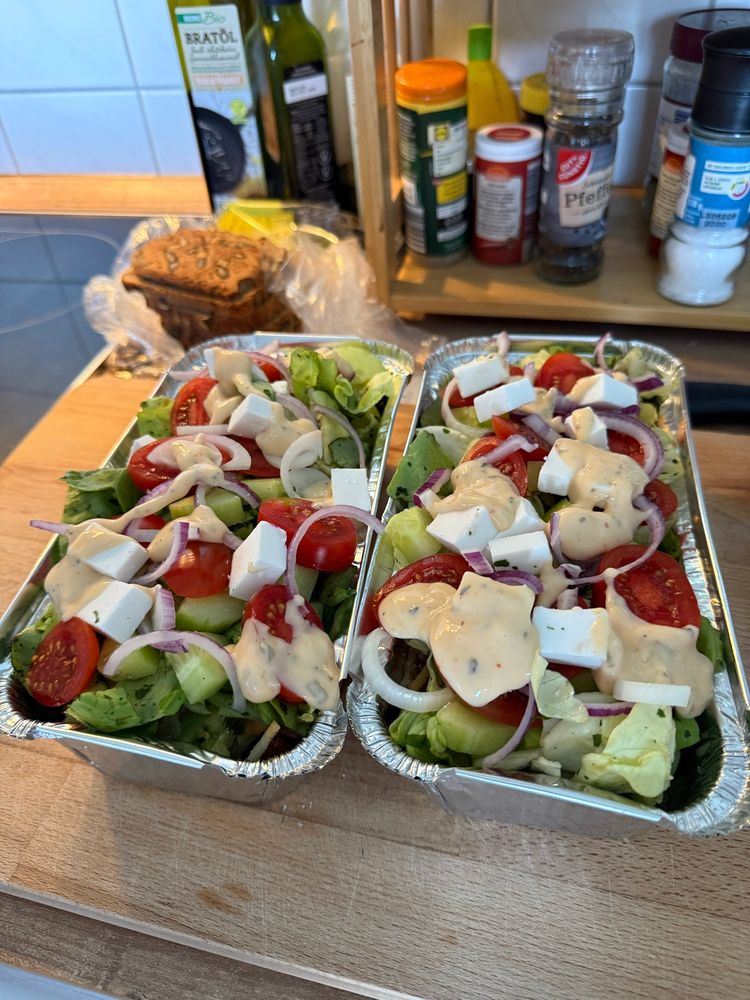 Zwei frische, vegane Salate in Aluschalen mit grünen Blattsalaten, Cherrytomaten, Gurken, roten Zwiebeln, veganem Feta und einem cremigen Dressing. Im Hintergrund sind verschiedene Gewürze und Öle zu sehen.