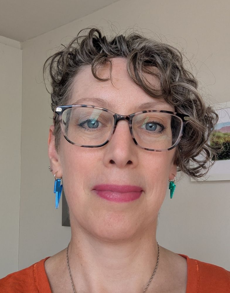 A white-passing woman is wearing a pair of shiny earrings, one blue and one green, in the shape of thunderbolts. She has blue eyes, glasses, pink lipstick and curly, brown hair cut in an asymmetrical pixie style. 