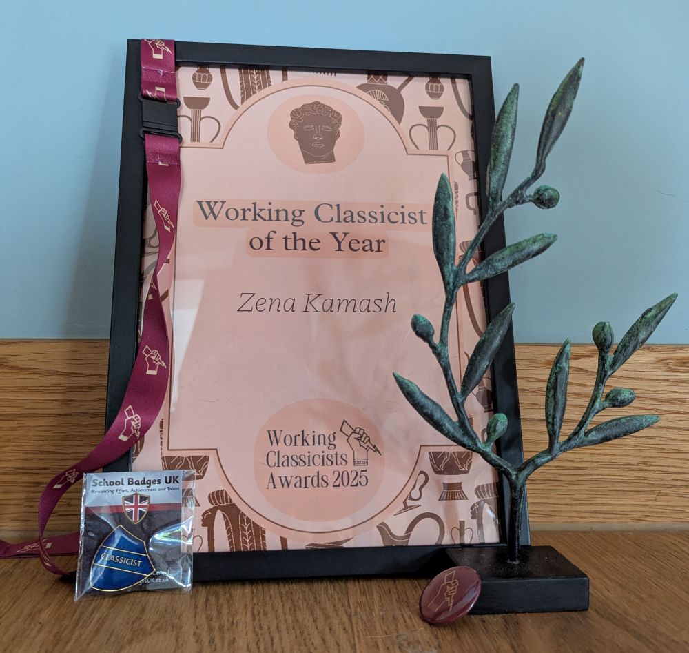 A framed certificate reads "Working Classicist of the Year Zena Kamash Working Classicists Awards 2025"; the certificate is beige with a border of Greco-Roman pottery in brown. Hanging on the top left corner of the frame is a burgundy lanyard, which is decorated in gold with the Working Classicists logo of a closed fist holding a thunderbolt. By the bottom left corner is a blue pin badge in the shape of a shield that reads "Classicist" in gold. In front of the frame to the right is an olive branch sculpture on a black rectangular base. Propped up against the base is a circular, burgundy badge with the Working Classicists logo in gold. All of these are against a background of oak at the bottom and a painted duck egg blue wall above.