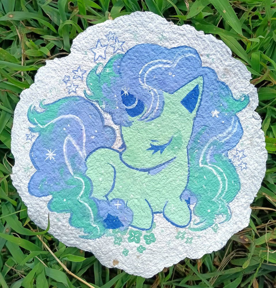 photo of a gouache painting laying in the grass. it depicts a galarian ponyta in pastel tones, surrounded by sparkles and four-leafed clover. the painting is on a circle of handmade paper about 7" across.