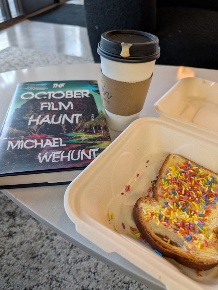 A hardcover of Michael Wehunt's novel THE OCTOBER FILM HAUNT sits next to a white takeout cup and a cardboard takeout box open to show fairy bread, its sprinkles bright. The red and yellow sprinkles pick up colors from the book cover.