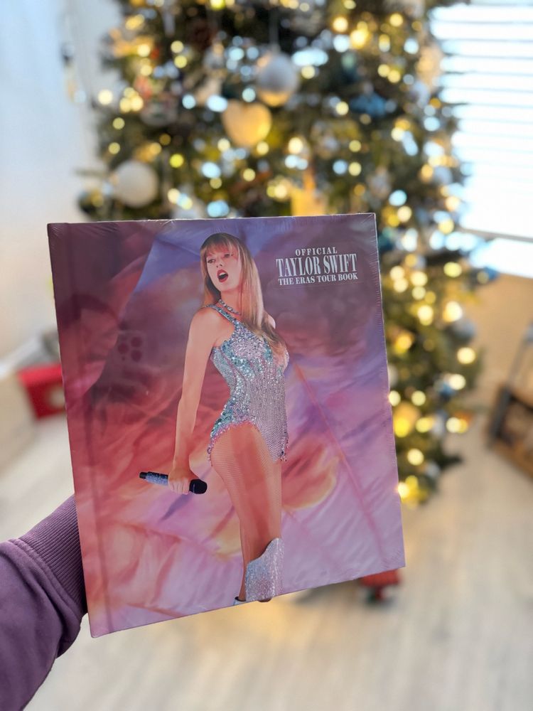 A photo of Taylor Swift’s Eras tour book in front of a Christmas tree. The book cover has Taylor standing on stage in a pink blue and silver body suit while holding a mic