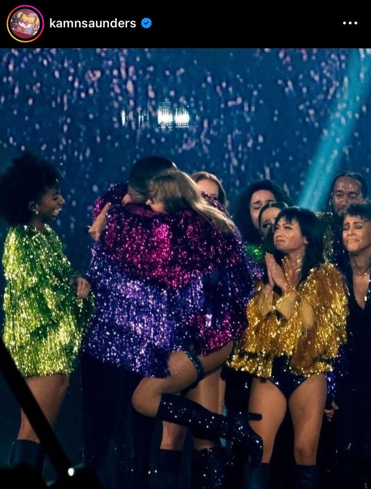 kamnsaunders post on Instagram, Taylor Swift wears a magenta fringe jacket and glitter knee high boots while hugging Kam Saunders who is in a purple fringe jacket and dark pants. Other dancers surround them smiling wearing their own fringe jackets in green, yellow.