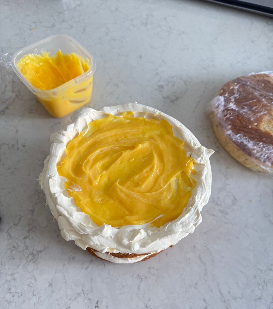 Mid-filing a cake process: A layer of cake with a buttercream border and lemon curd filling. Another sponge layer is in clingfilm in the right background and a tub of lemon curd in the left background. 