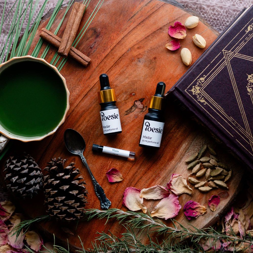 A wooden table with small oil perfume bottles, a cup of green liquid (likely matcha), pine cones, a book, and dried botanicals. The bottles are labeled "Poesie", "Madar".