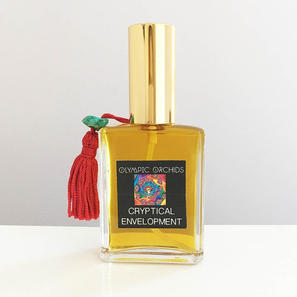 a square-rectangular perfume bottle with a gold cylindrical lid and a red tassel with a green stone attached hanging from the cap. the label is black with white text. the text reads "Olympic Orchids", "Cryptical Envelopment". In between the 2 pieces of text there is a small picture of rainbow-colored, psychedelic concentric swirls.