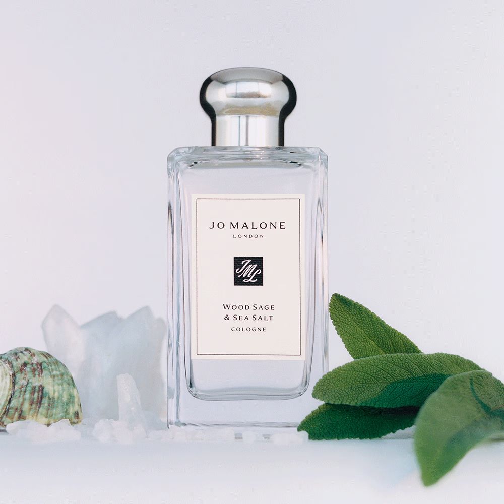 A square-base, rectangular clear bottle of perfume with a silver cap that looks like a lightbulb with a wide base and flat top. it's on a white background, with sage leaves to the right and salt crystals and a green and brown seashell to the left. the label is off-white, and reads from top-down "Jo Malone", "London", *LOGO*, "Wood Sage & Sea Salt", "Cologne".