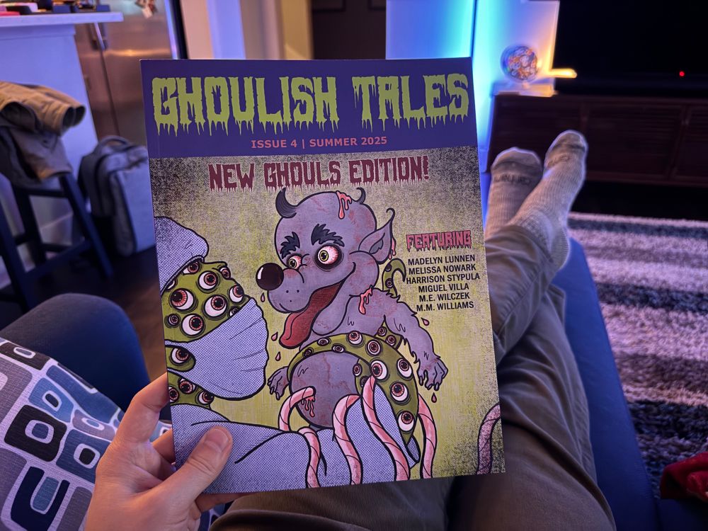 Holding a copy of Golish Tales: issue four - new ghouls edition while sitting on a blue sectional in green jeans and grey socks. 