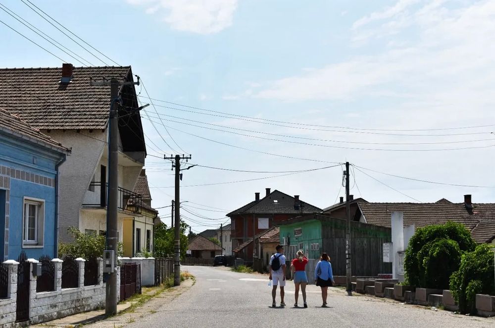 Eine ruhige, fast menschenleere Dorfstraße in einem ländlichen Gebiet. Es ist Tag, der Himmel ist hell und leicht bewölkt. Links und rechts der Straße stehen ältere Häuser mit Ziegeldächern, manche farbenfroh gestrichen – zum Beispiel ein auffällig blaues Haus im Vordergrund. Einige Häuser wirken verlassen oder wenig gepflegt. Stromleitungen verlaufen in mehreren Richtungen über die Straße hinweg.

In der Mitte des Bildes, mit dem Rücken zur Kamera, gehen drei junge Menschen nebeneinander auf der Straße. Sie tragen sommerliche Kleidung – kurze Hosen, T-Shirts, Rucksäcke. Die Straße ist asphaltiert, aber leer, ohne Autos oder andere Menschen. Die Szene wirkt still, beinahe verlassen, was den Eindruck eines strukturschwachen oder entvölkerten Dorfes verstärkt.