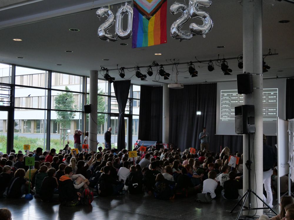 Aula einer Schule. Der Raum ist modern, mit großen Fenstern an der linken Seite, durch die Tageslicht einfällt. Draußen sieht man Bäume und ein Schulgebäude. Im Vordergrund sitzen viele Schüler:innen auf dem Boden, in Gruppen nach Klassen sortiert – sichtbar an den hochgehaltenen Schildern mit Klassenbezeichnungen wie „7a“, „7b“, „8a“ usw. Sie sind überwiegend in Freizeitkleidung und tragen teilweise Rucksäcke. Einige Kinder heben die Hand, als Reaktion auf eine Frage oder Abstimmung. Vorne rechts auf einer Bühne steht eine Person mit Mikrofon und spricht, während hinter ihr eine große Leinwand hängt. Auf dieser Leinwand ist ein Stunden- oder Raumplan zu sehen, mit verschiedenen Sessions oder Workshops. Über der Bühne hängen silberne Ballons mit der Zahl „2025“. Darüber befindet sich eine große Pride-Flagge mit einem zusätzlichen farbigen Pfeilmuster (Progress-Pride-Flagge), die Vielfalt und Inklusion symbolisiert. Die Stimmung wirkt lebendig und organisiert.