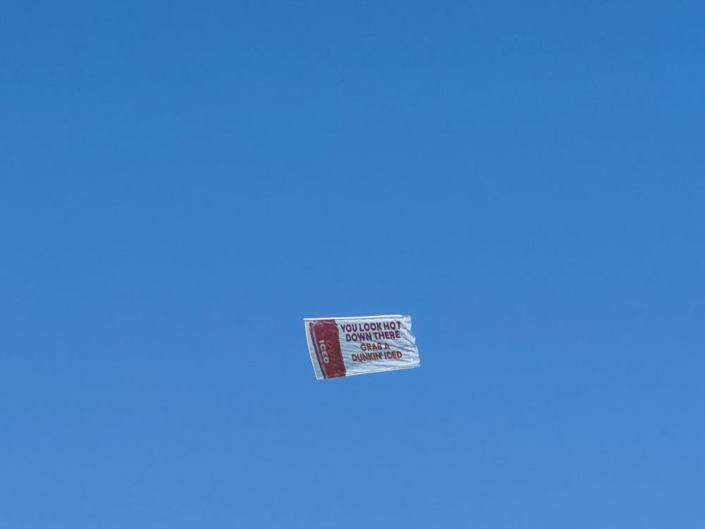 Over a clear blue sky, a banner pulled by an airplane along a beach reads "YOU LOOK HOT DOWN THERE / GRAB A DUNKIN' ICED"