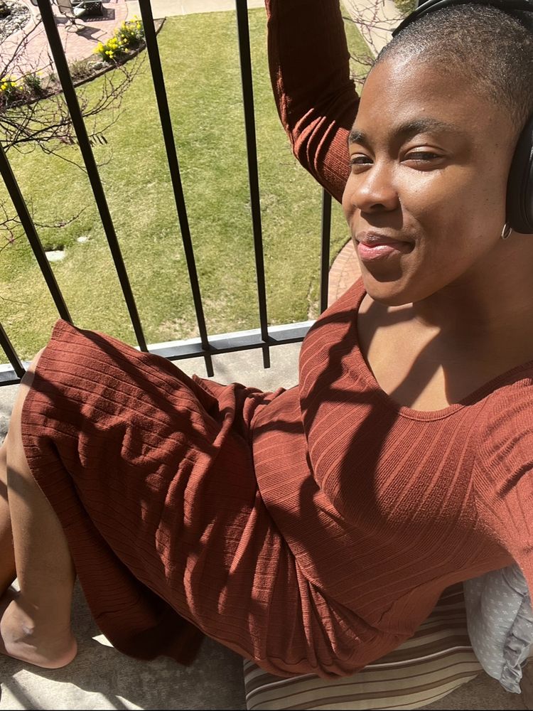 A Black nonbinary person wearing a orangish brown dress and their noise cancelling headphones on their patio with the sun shining on them.