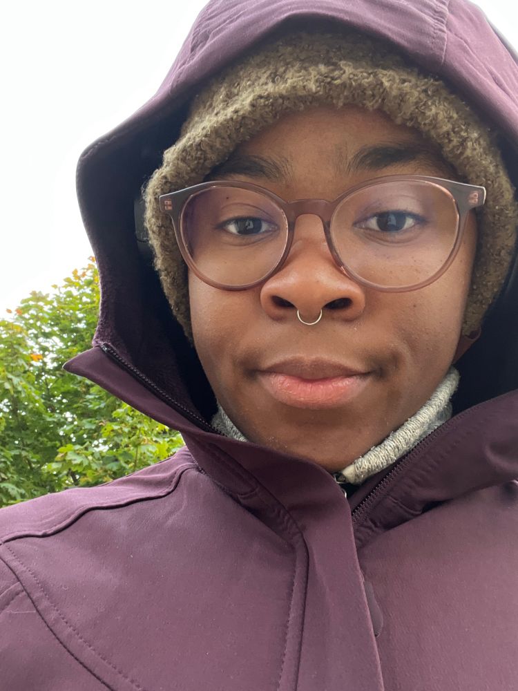 Me outside on a mental health walk in a portrait like selfie, a Black nonbinary person with glasses a septum ring, a green hat and a purple jacket with the hood up.