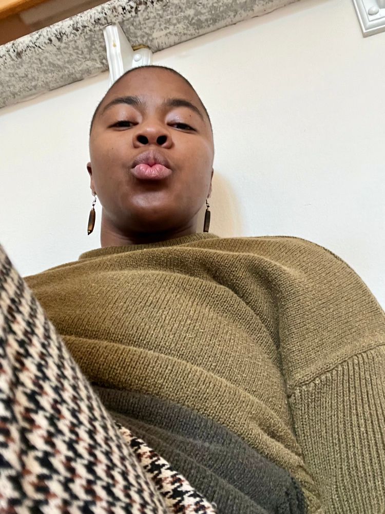 Black nonbinary person w/ a buzzed haircut looking down into the camera.