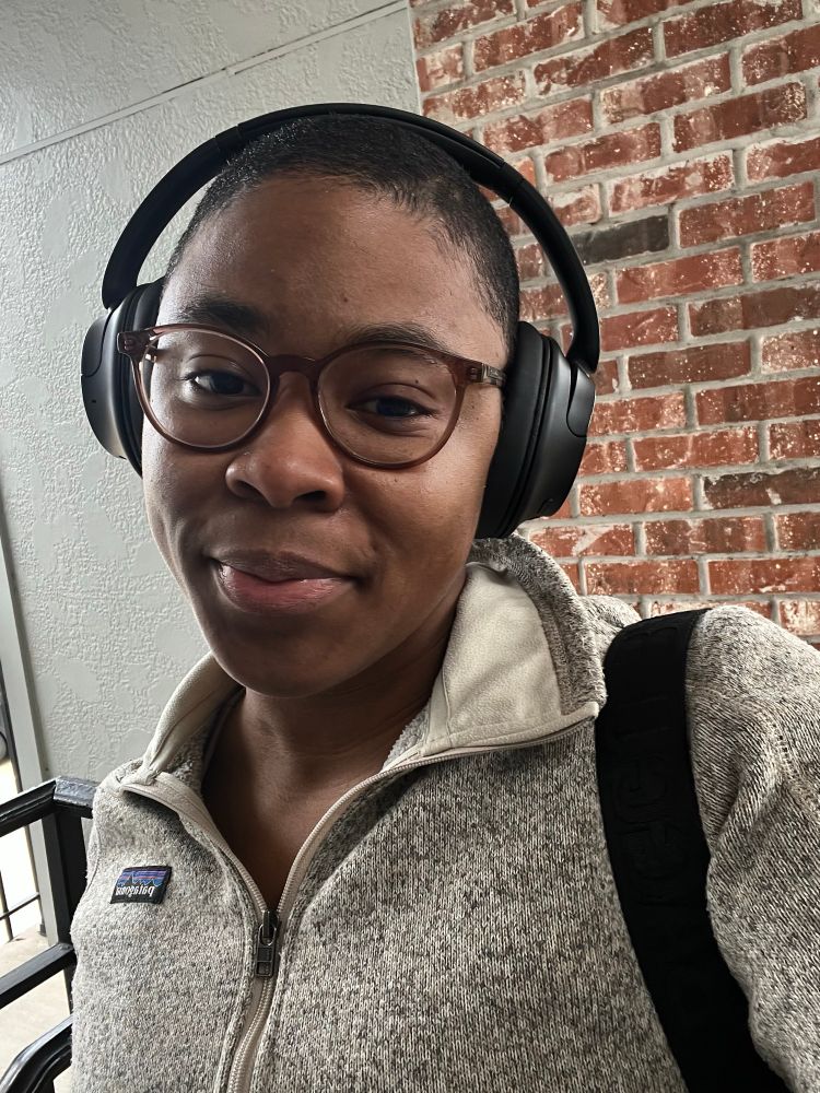 Brown-skinned Black person with a buzzed head and glasses, looking into the camera kindly.